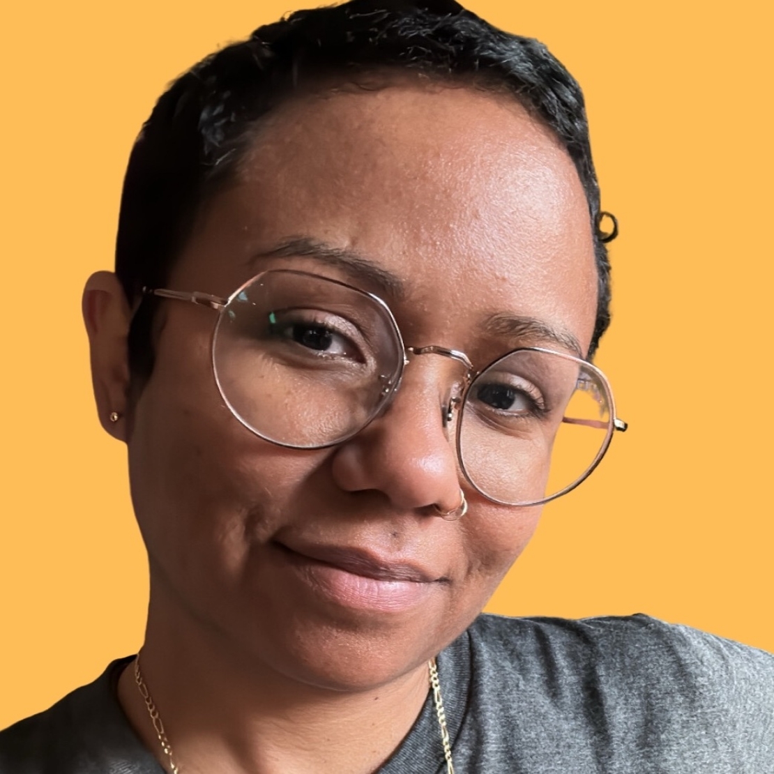 A headshot of Sharon in a gray T_shirt and wearing a gold chain necklace and glasses.